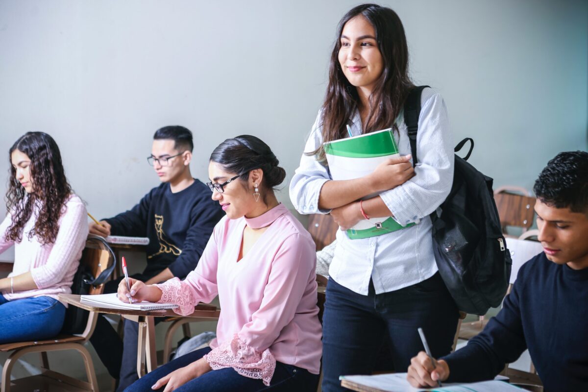 教室でノートを取る学生たちの中、ノートを抱えながら立ち上がる女性学生。