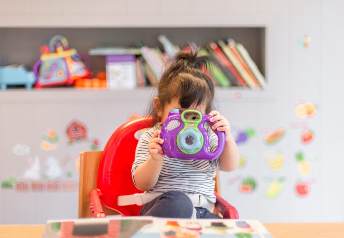 カラフルなおもちゃのカメラを手に持つ幼い子ども。背景には本棚と教育用のおもちゃが配置されている。