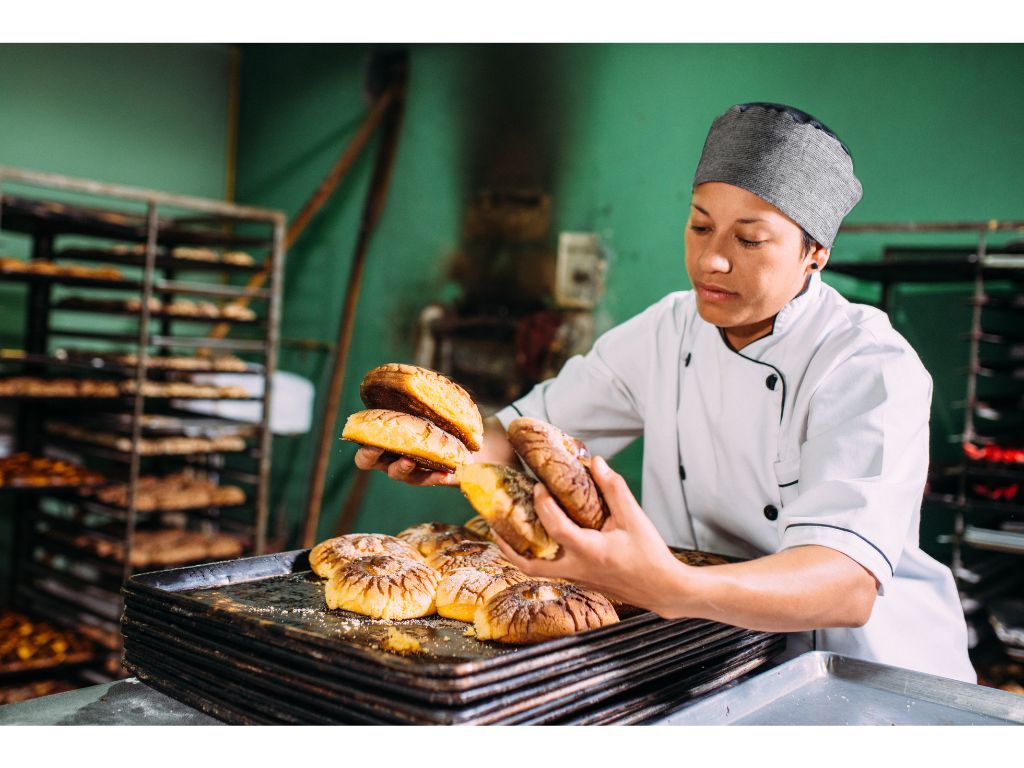 パン職人が焼き上がったパンをトレイに丁寧に並べている様子。背景にはパンが置かれたラックが見える。