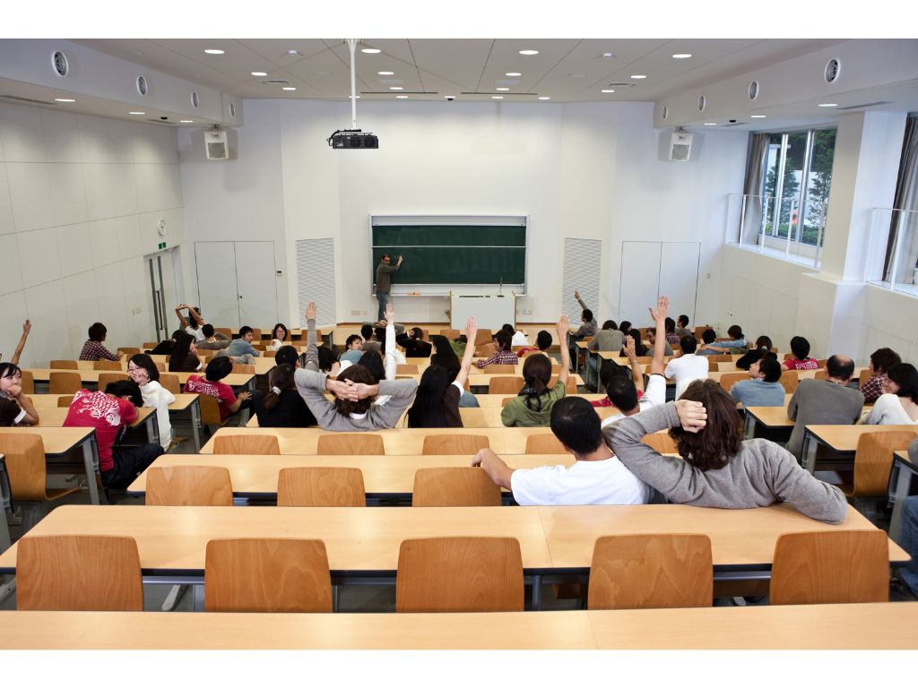 大学の講義室で、学生たちが講義を受けている様子。数人が手を挙げ、講師が黒板の前に立っている。